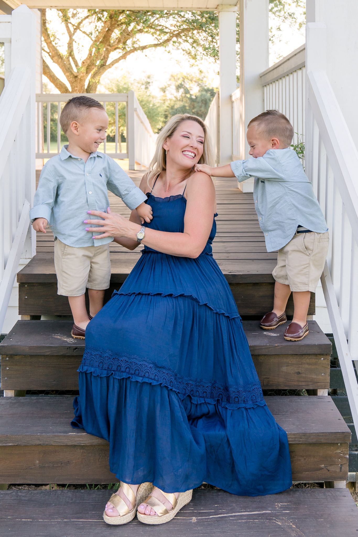 A smiling mother in a blue maxi dress sits on wooden steps with her two young sons in matching blue shirts.