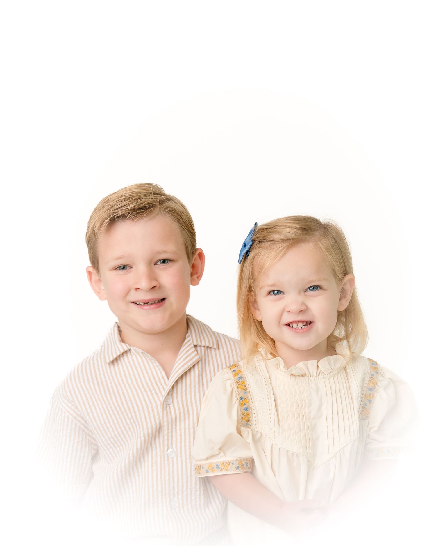 Smiling blonde boy in striped shirt poses with younger blonde toddler sister in cream smocked dress.