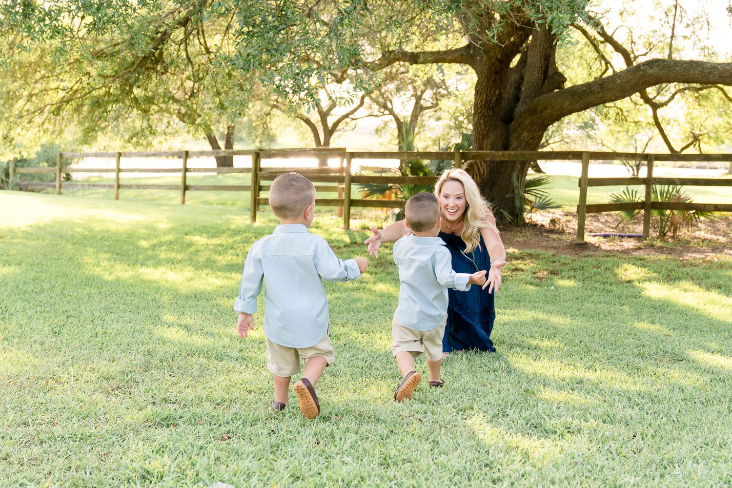 A joyful blonde mother kneels in a sunny green field as her two young sons run toward her.