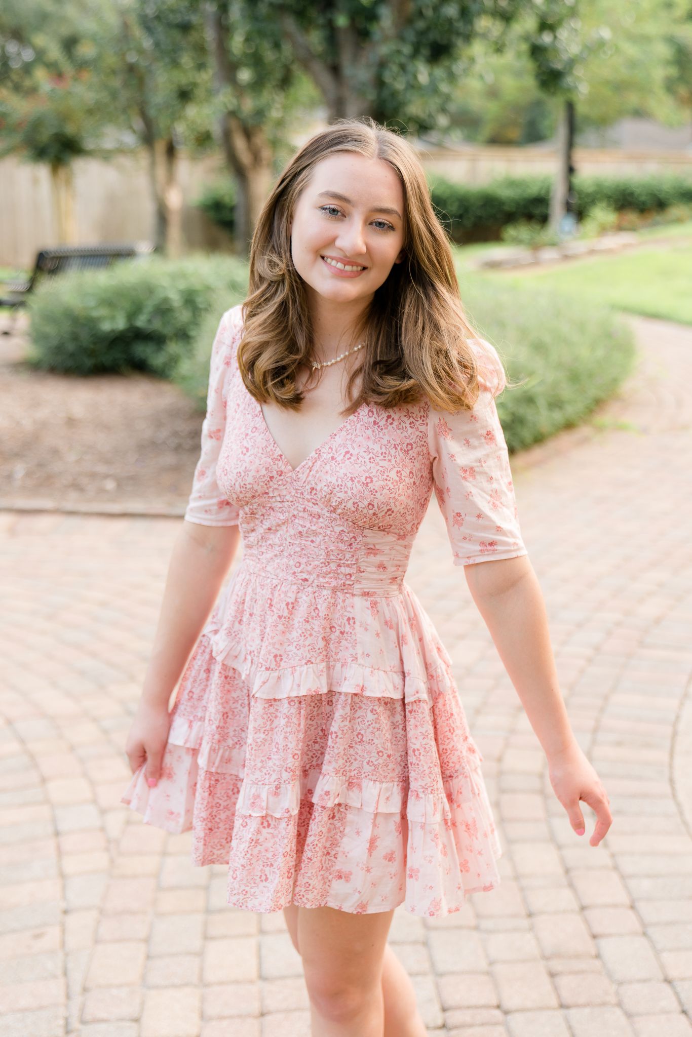 Young woman smiling in a pink floral tiered mini dress, posing outdoors on a brick pathway surrounded by greenery.