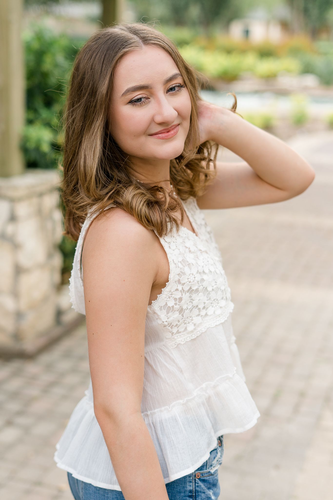 Young woman in white lace top and jeans smiling outdoors near stone columns in a garden setting.