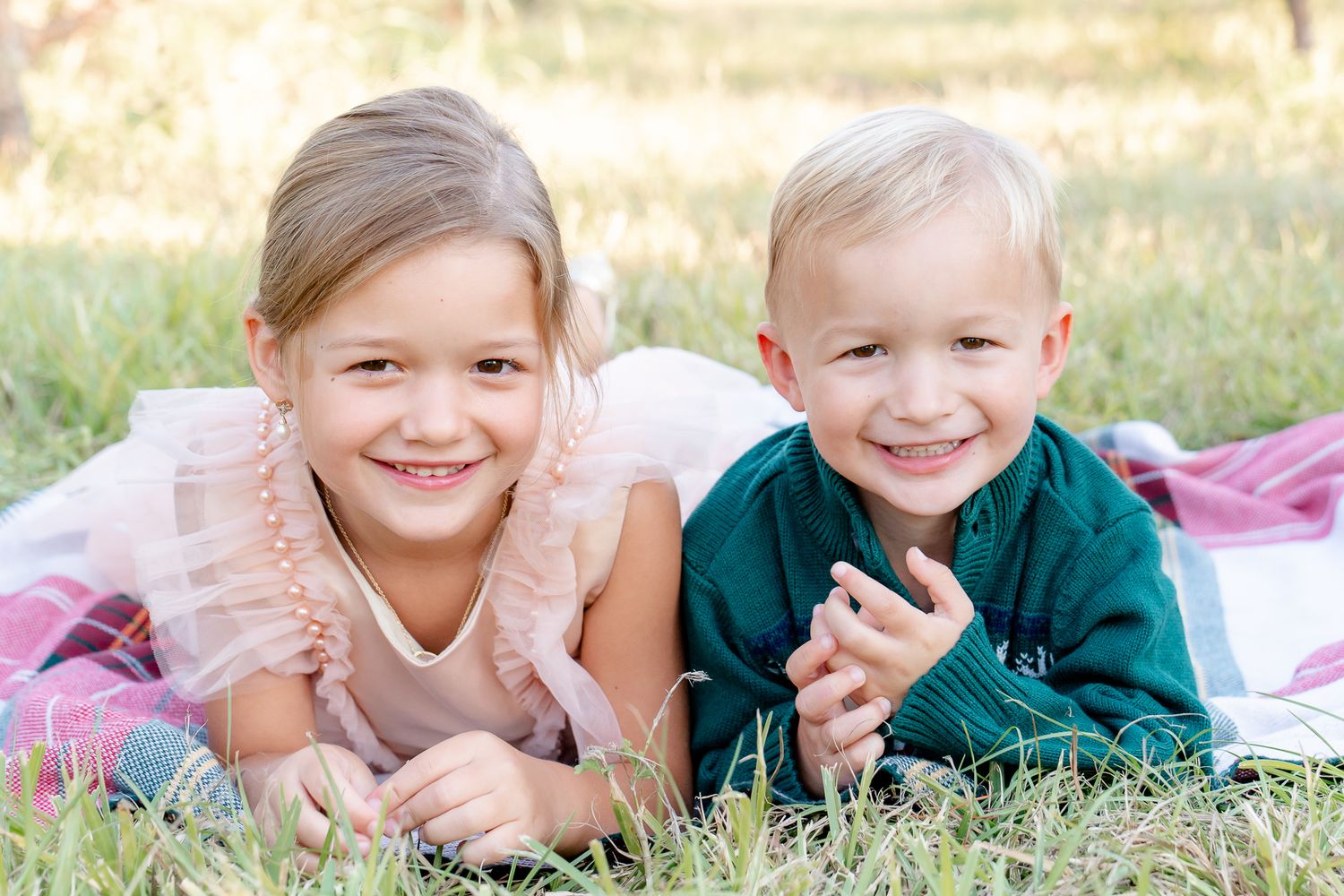 Two smiling blonde siblings lying on a pink blanket in the grass for an outdoor portrait.
