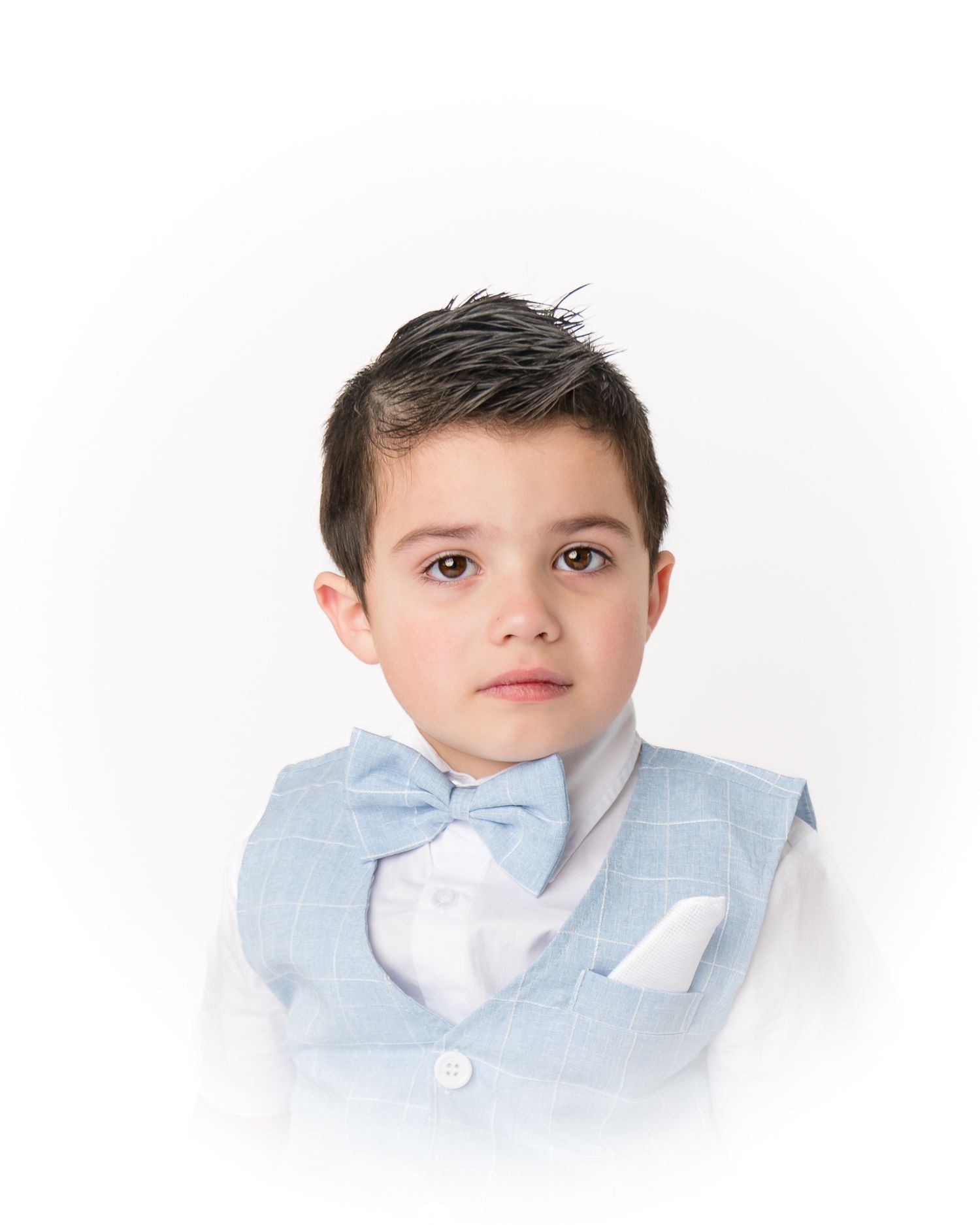 A young boy with dark hair poses seriously in a light blue vest, bow tie, and white dress shirt.
