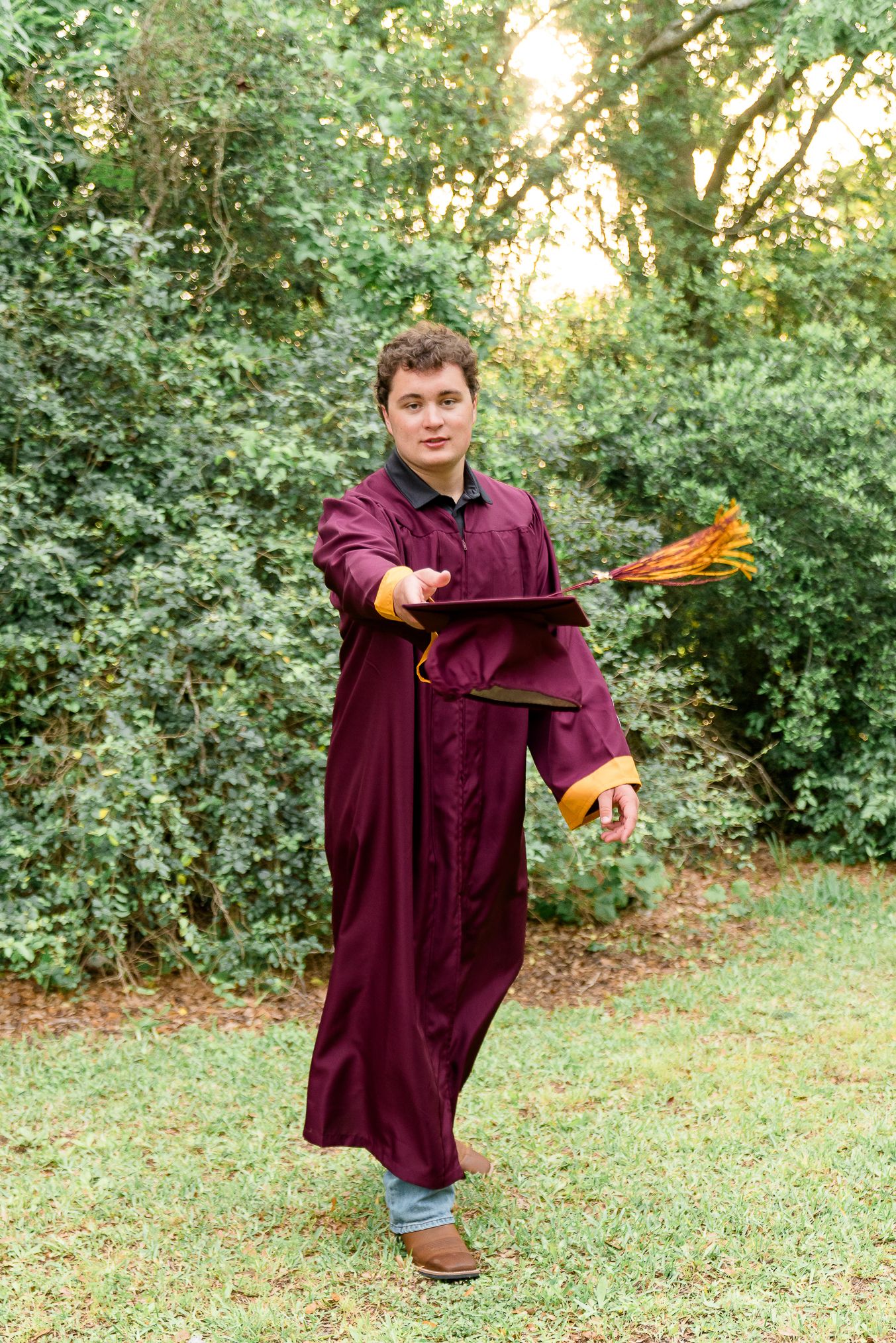 Young man in maroon graduation gown holding cap outdoors among green trees on grass.