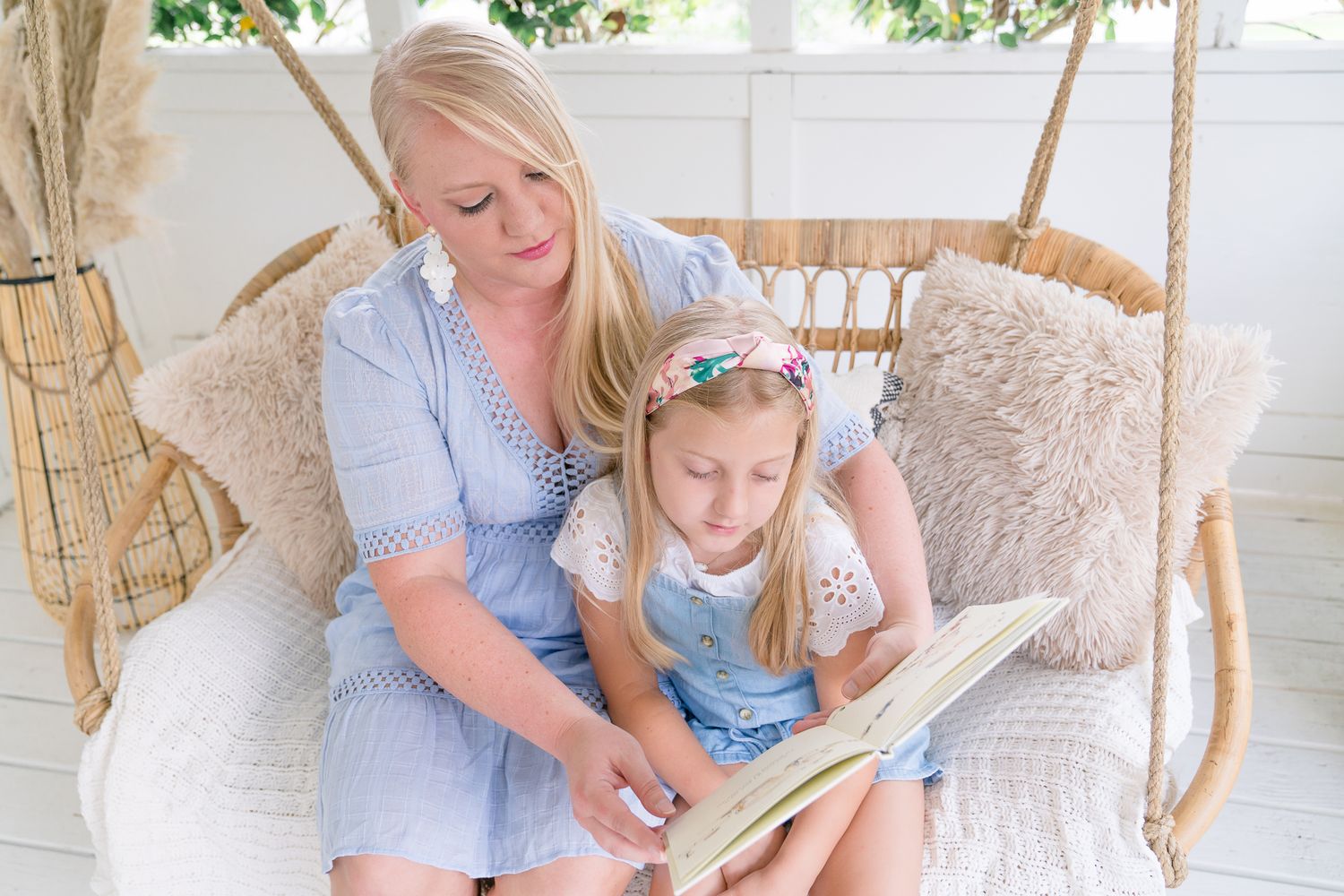 Blonde mother in blue dress reads a book with young blonde daughter on cozy rattan porch swing.