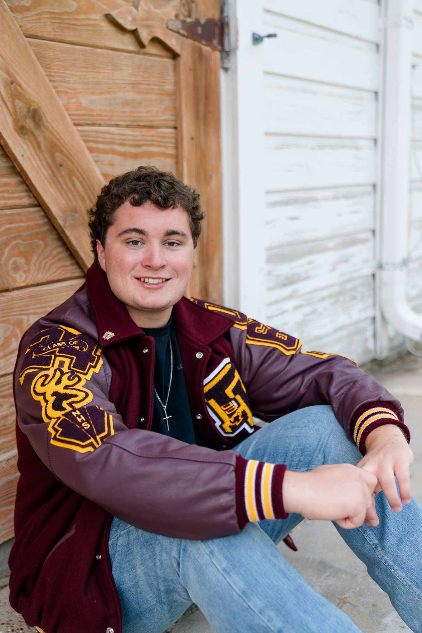 Smiling teenage boy in a maroon varsity letterman jacket sitting against a rustic wooden door for senior photos.