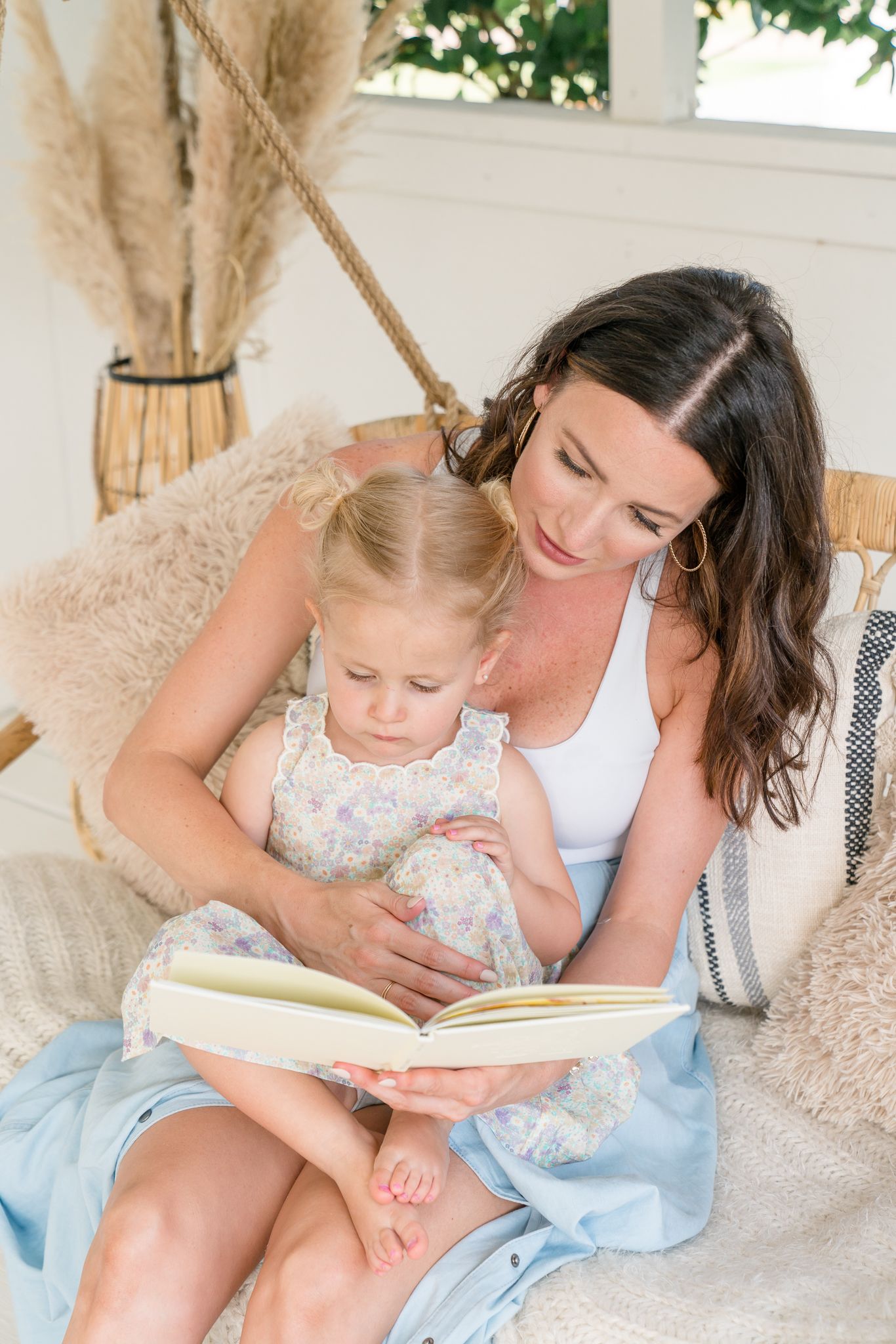 A dark-haired mother reads a book with her young blonde daughter on a cozy hanging swing seat.