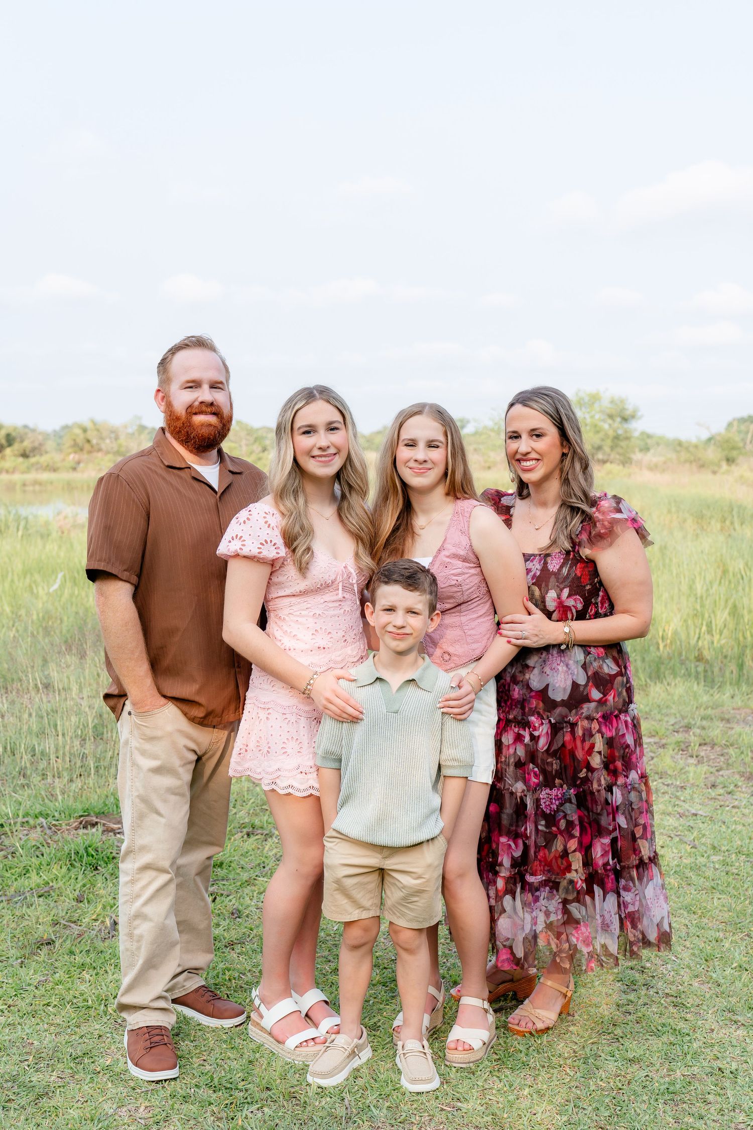 A family of five smiles together in a green outdoor field setting.