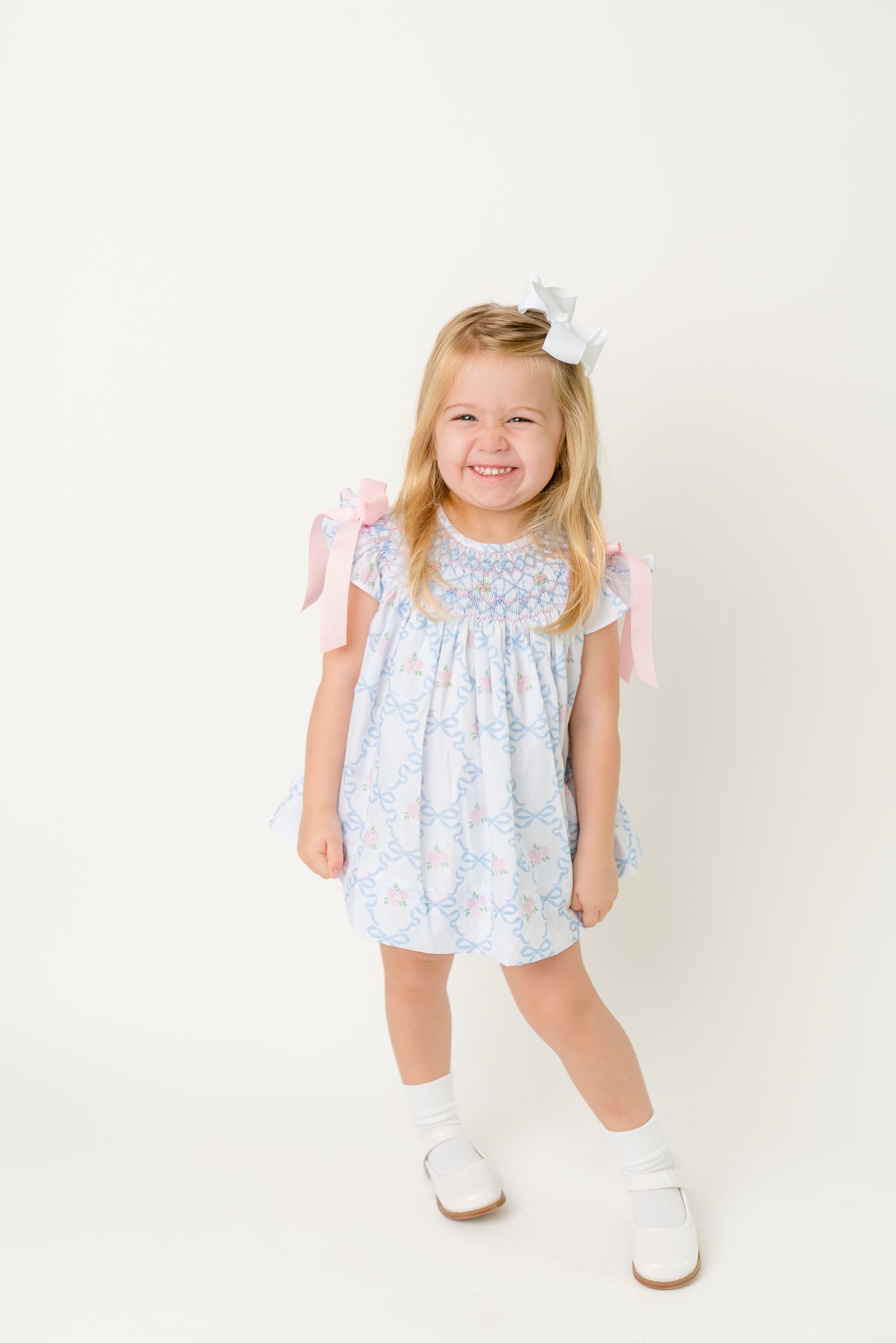 Smiling blonde toddler girl in blue floral smocked dress with pink bows and white hair bow poses happily.