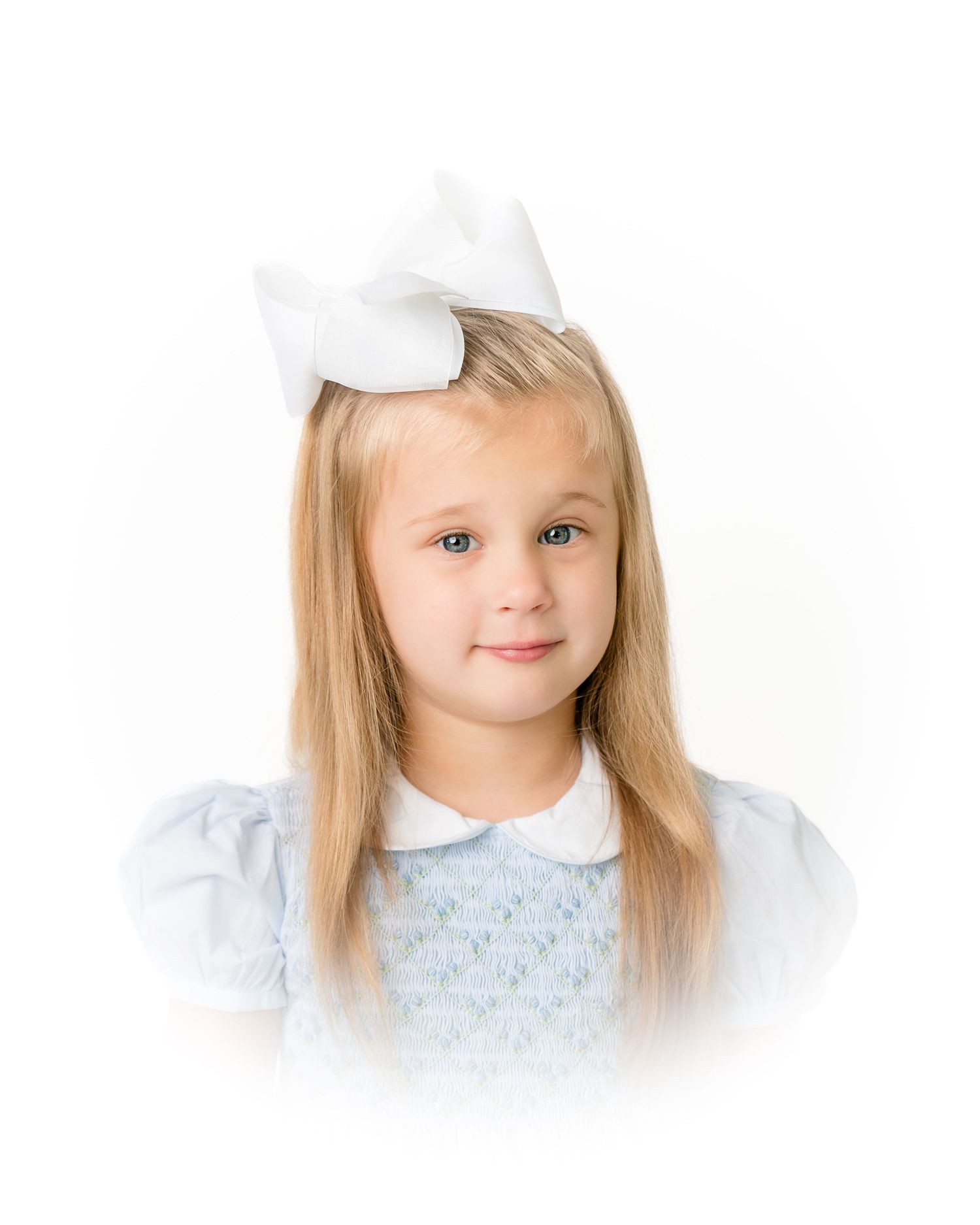 Young girl with long blonde hair wearing a white bow and light blue smocked dress portrait.