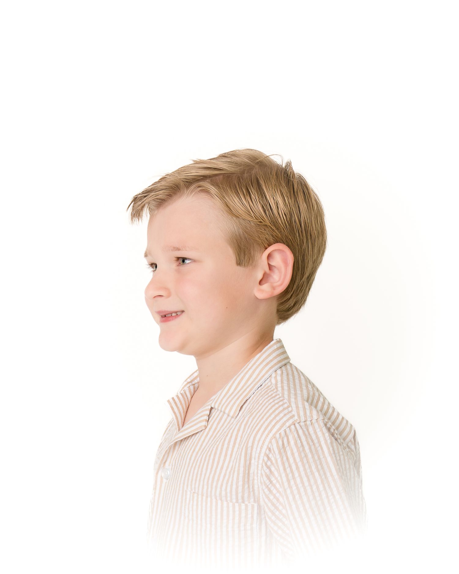 A young boy with blonde hair smiles in profile view, wearing a striped button-up shirt against a white background.