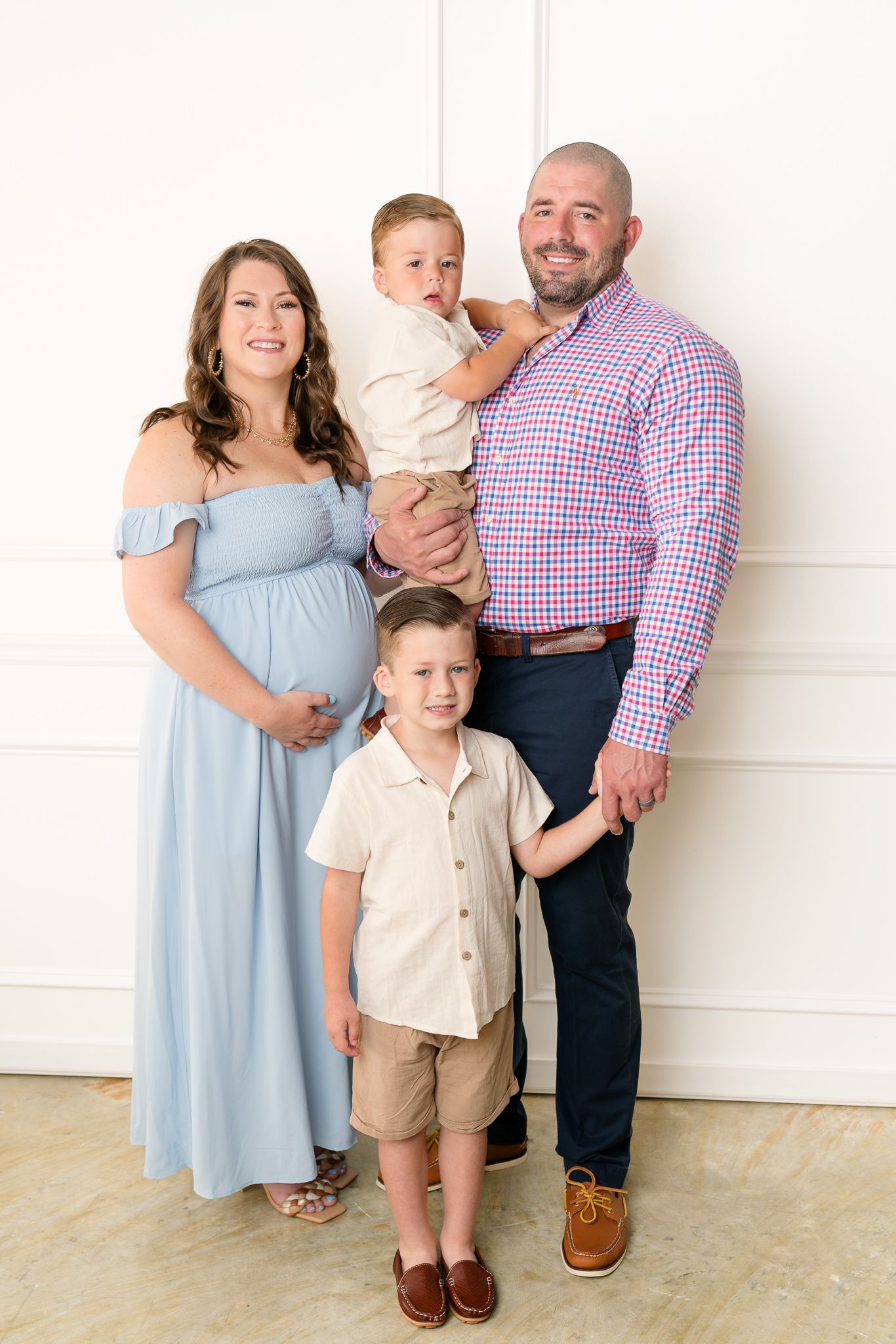 A pregnant mother in blue gown poses with her husband and two young sons for a studio family portrait.