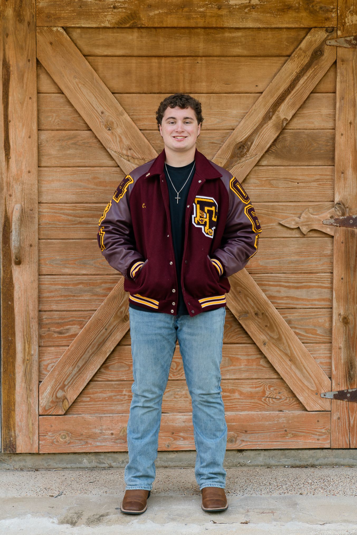 Teen boy in maroon letterman jacket and jeans smiling in front of rustic wooden barn doors.
