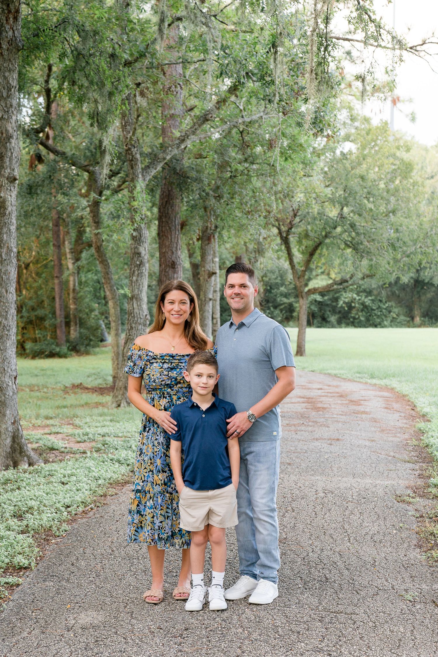 A family of three, parents and young son in navy, pose on a tree-lined gravel path outdoors.