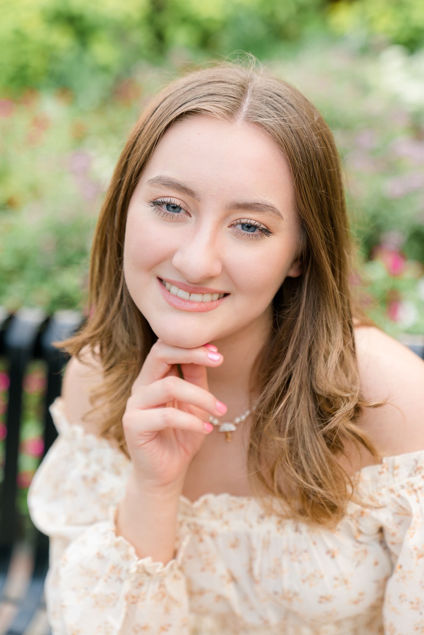 Young woman in floral off-shoulder dress smiling outdoors, hand on chin, with colorful garden flowers in background.