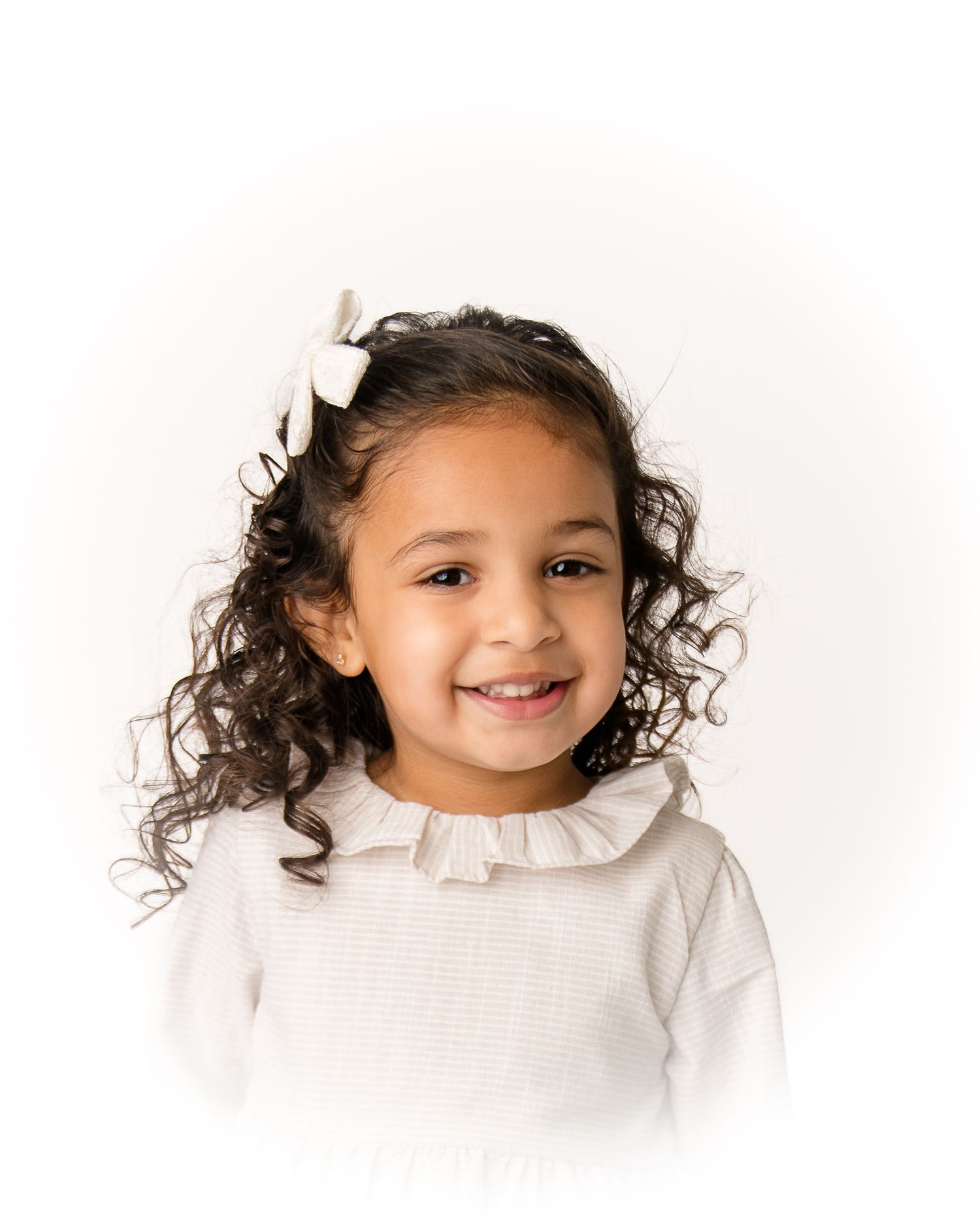 A cheerful young girl with curly dark hair smiles, wearing a white ruffled top and a small bow hair clip.