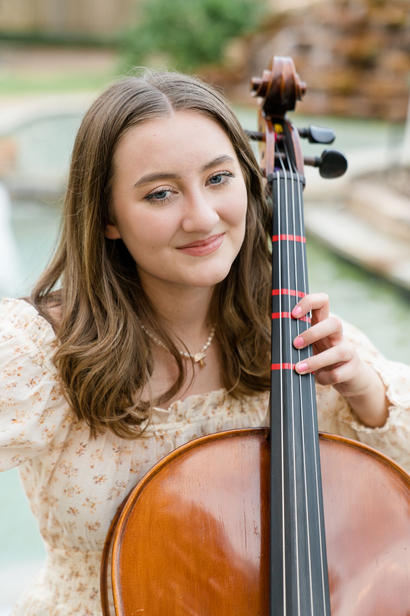 Young woman smiling while holding a cello outdoors near a fountain, wearing a floral dress and delicate necklace.