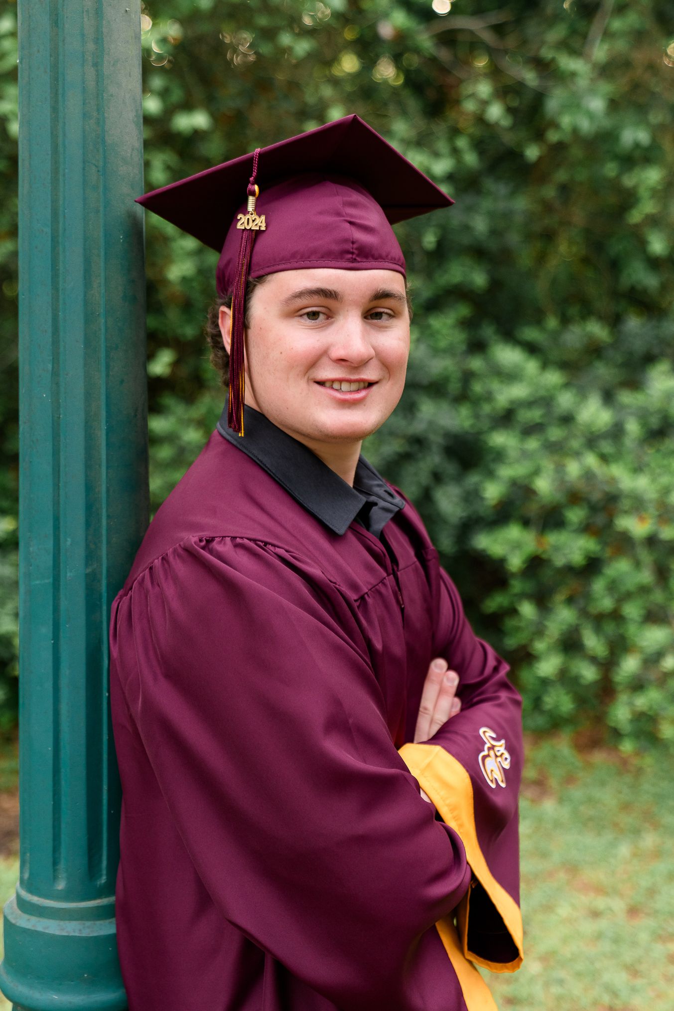 Smiling graduate in maroon cap and gown leaning against a green pole outdoors with lush greenery background.