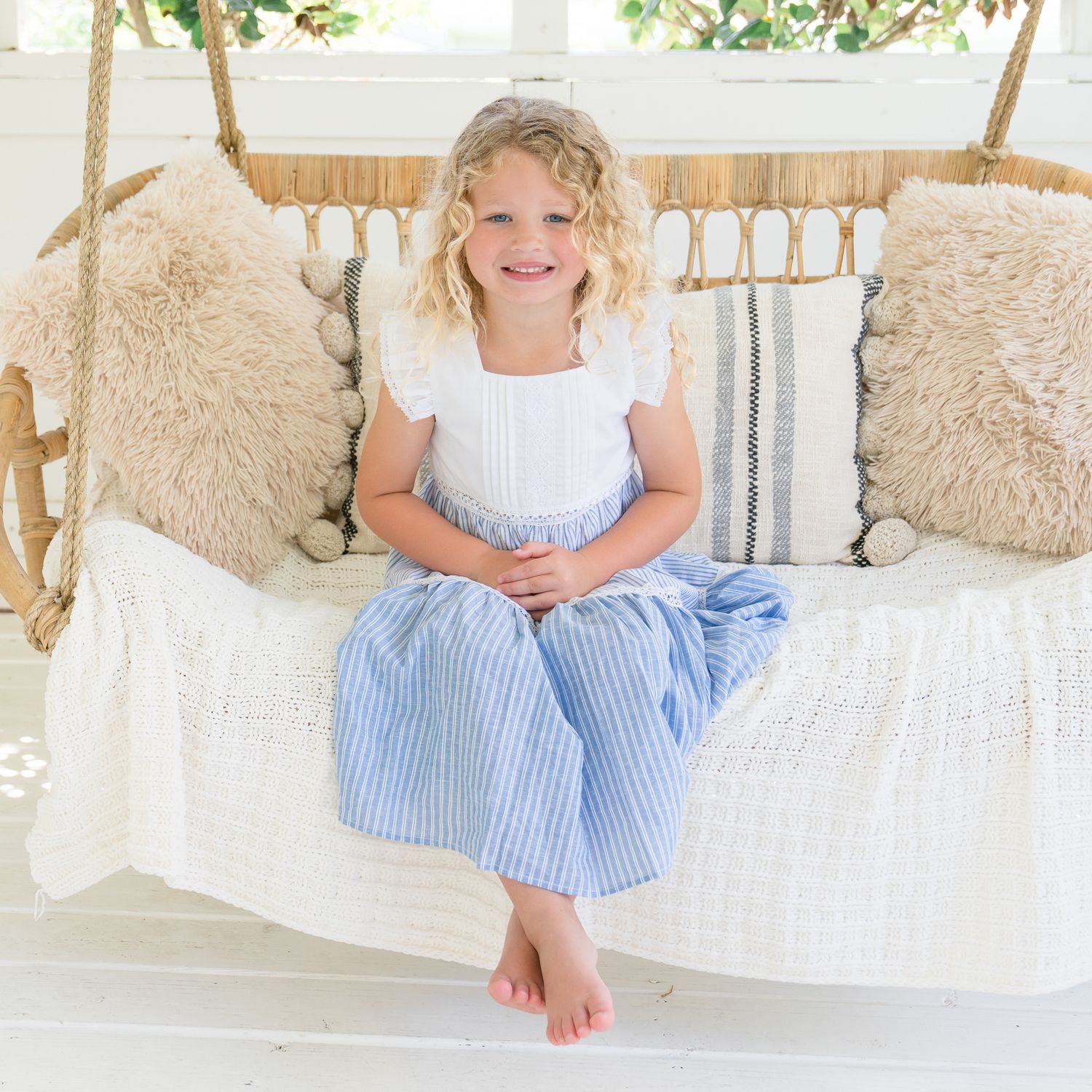 Blonde curly-haired girl in blue striped dress sitting barefoot on a boho hanging swing with cushions.