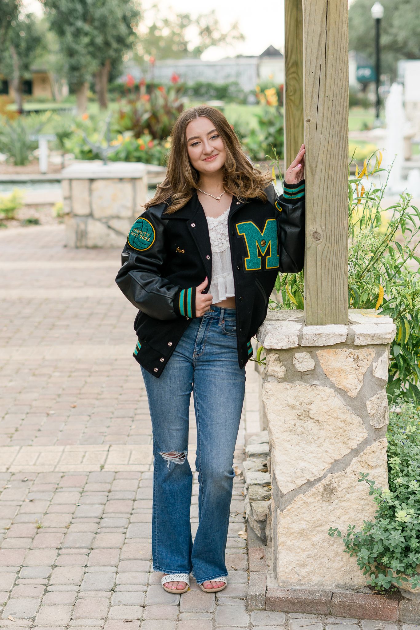 Teen girl in varsity jacket with M letter patch, ripped jeans, posing by stone column in garden.