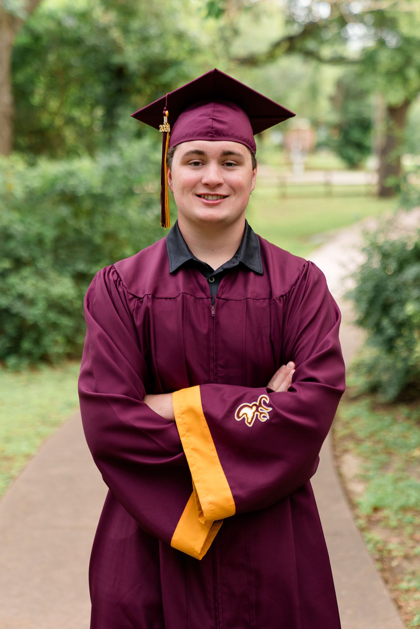 A smiling graduate in maroon cap and gown with gold accents stands arms crossed on a tree-lined pathway.