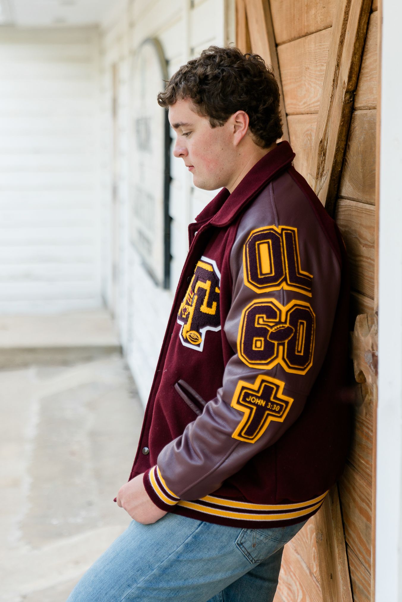 Teen boy in maroon varsity letterman jacket leaning against wooden wall, looking down, wearing jeans.