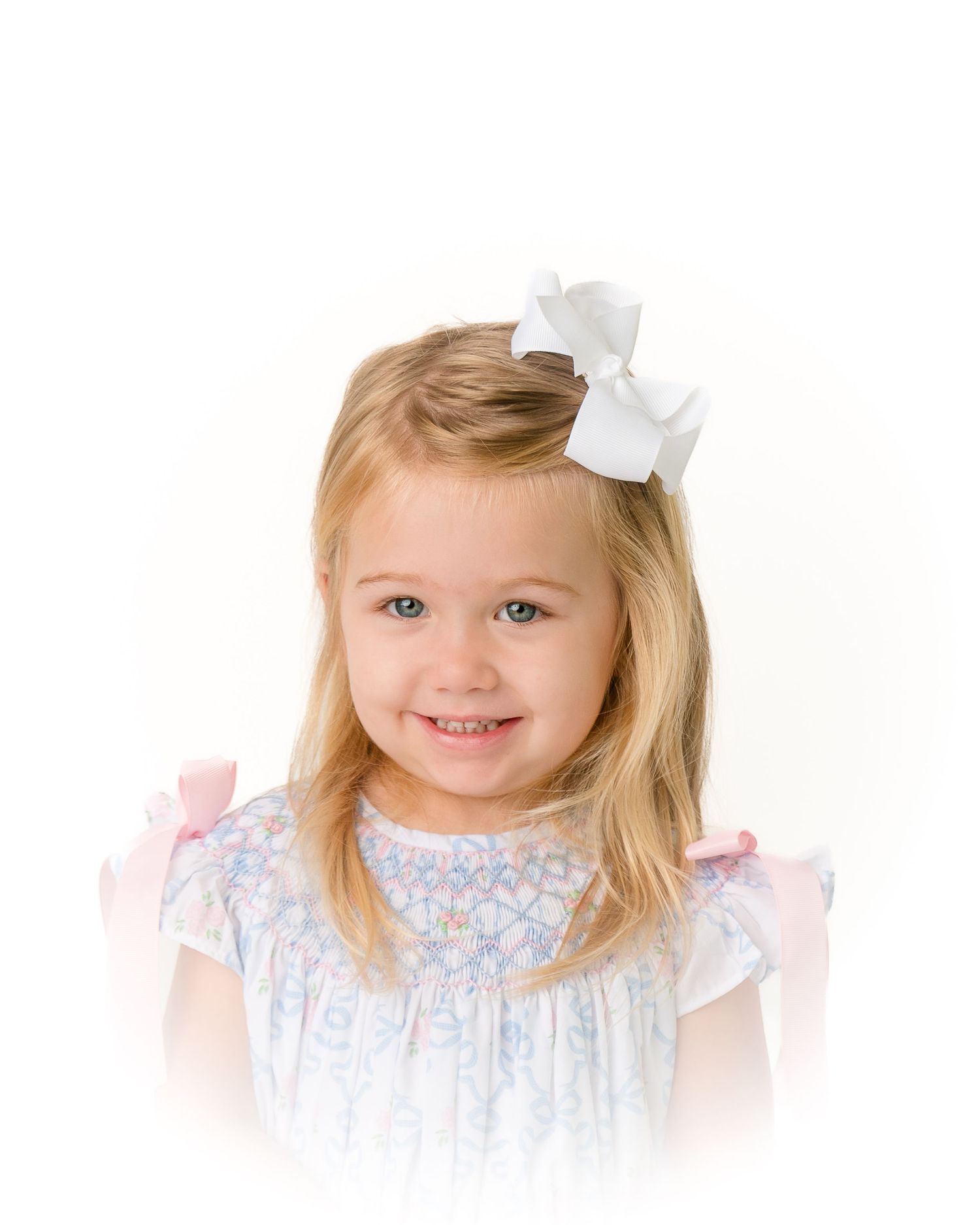 A smiling young girl with blonde hair wears a white bow and a light blue smocked dress with pink accents.