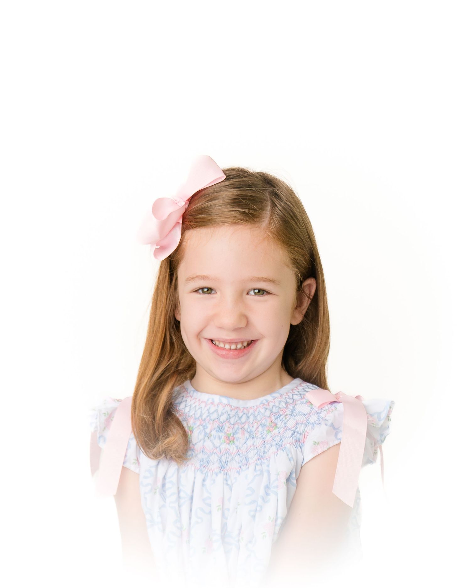 Happy young girl with brown hair and pink bow smiling in floral smocked dress.