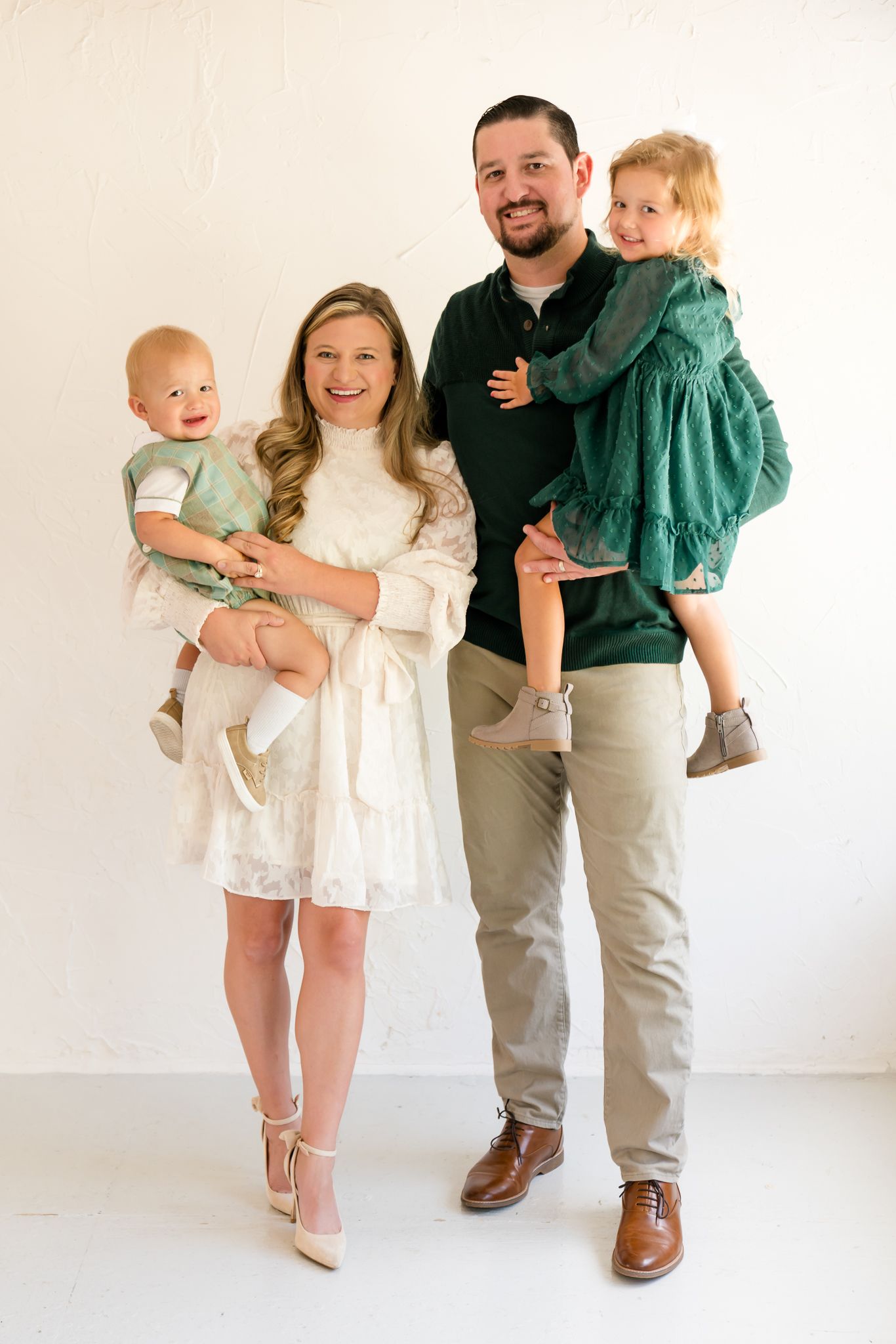 Smiling parents each hold a young child dressed in green and white during an indoor family portrait.
