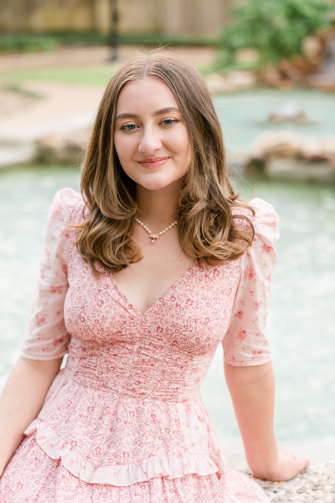 Young woman in pink floral dress with puff sleeves and pearl necklace smiling outdoors near a fountain.