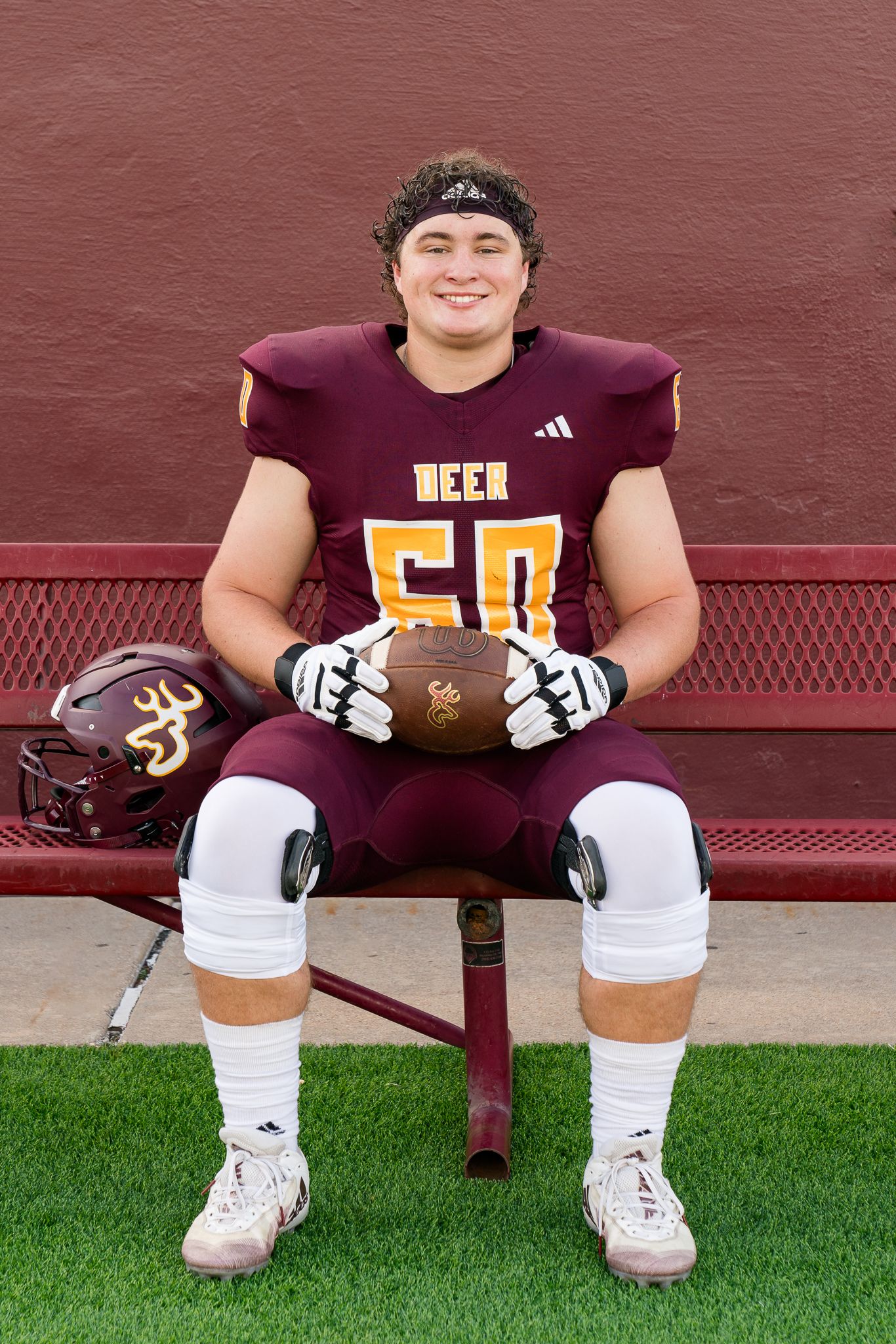 High school football player #50 in maroon Deer uniform sits on bench holding football during team photo day.