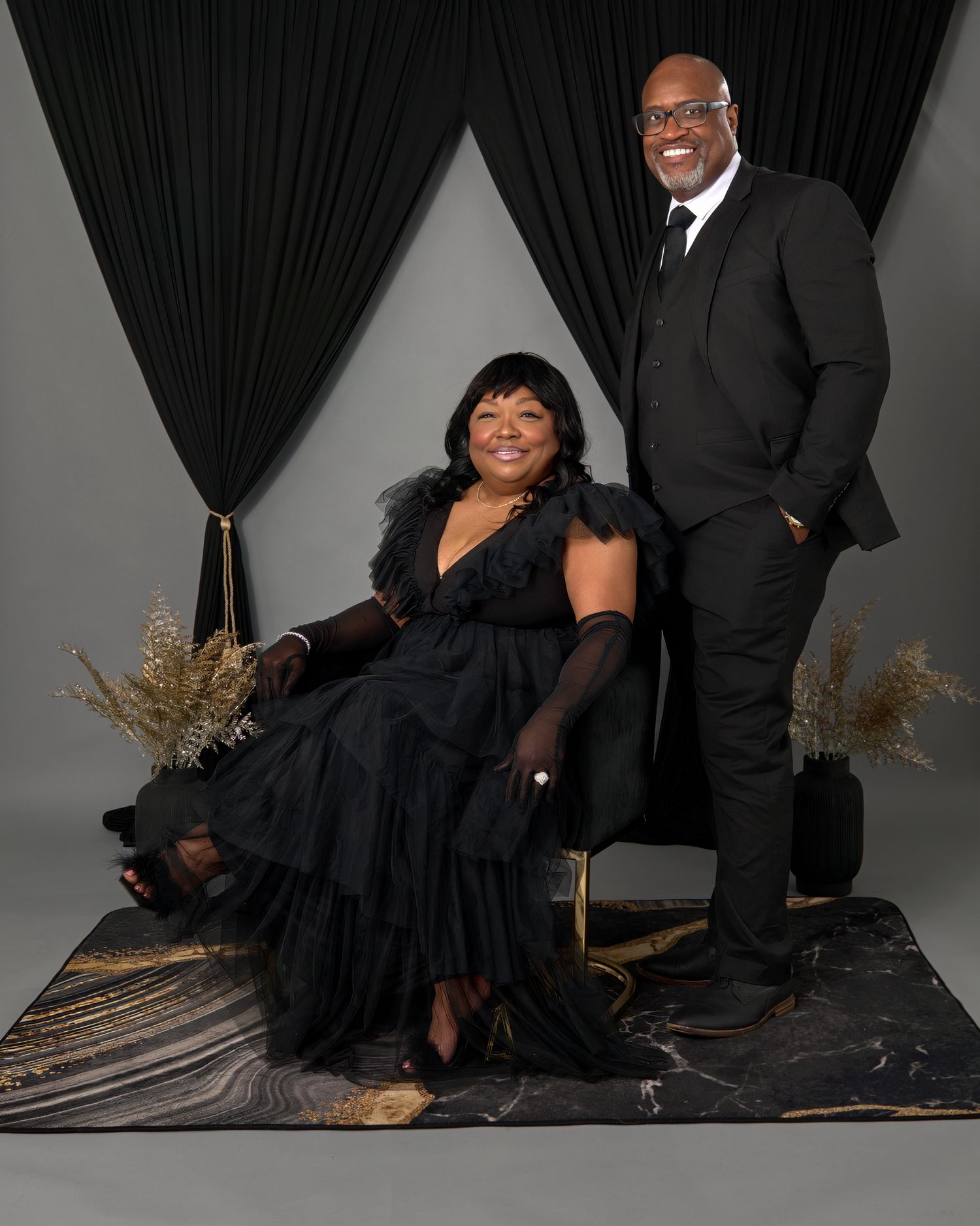 Elegant studio portrait with two people in formal black attire against a dark curtained backdrop.