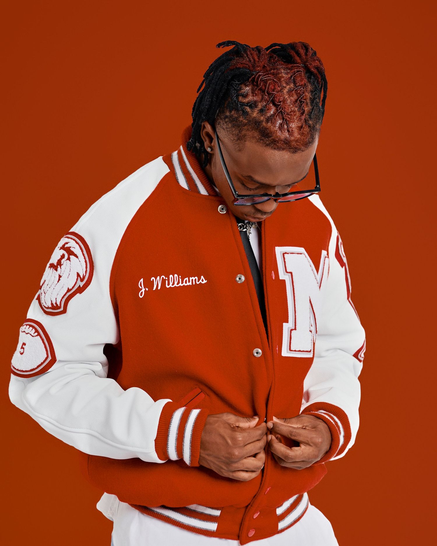 Person wearing red and white varsity jacket with J. Williams embroidery and braided hair against orange background.