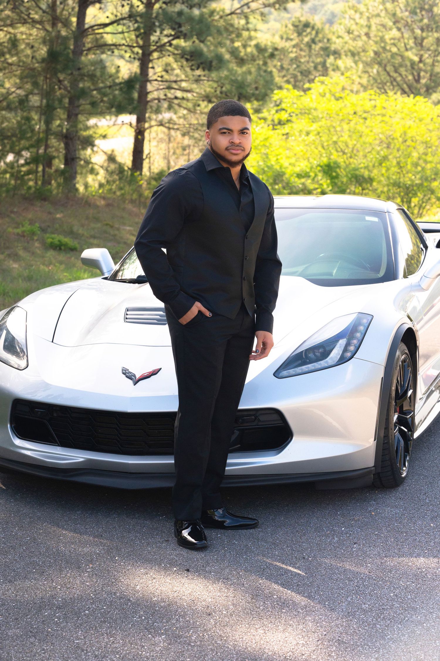 A person in all black attire stands confidently next to a sleek white Chevrolet Corvette on a scenic forest road.