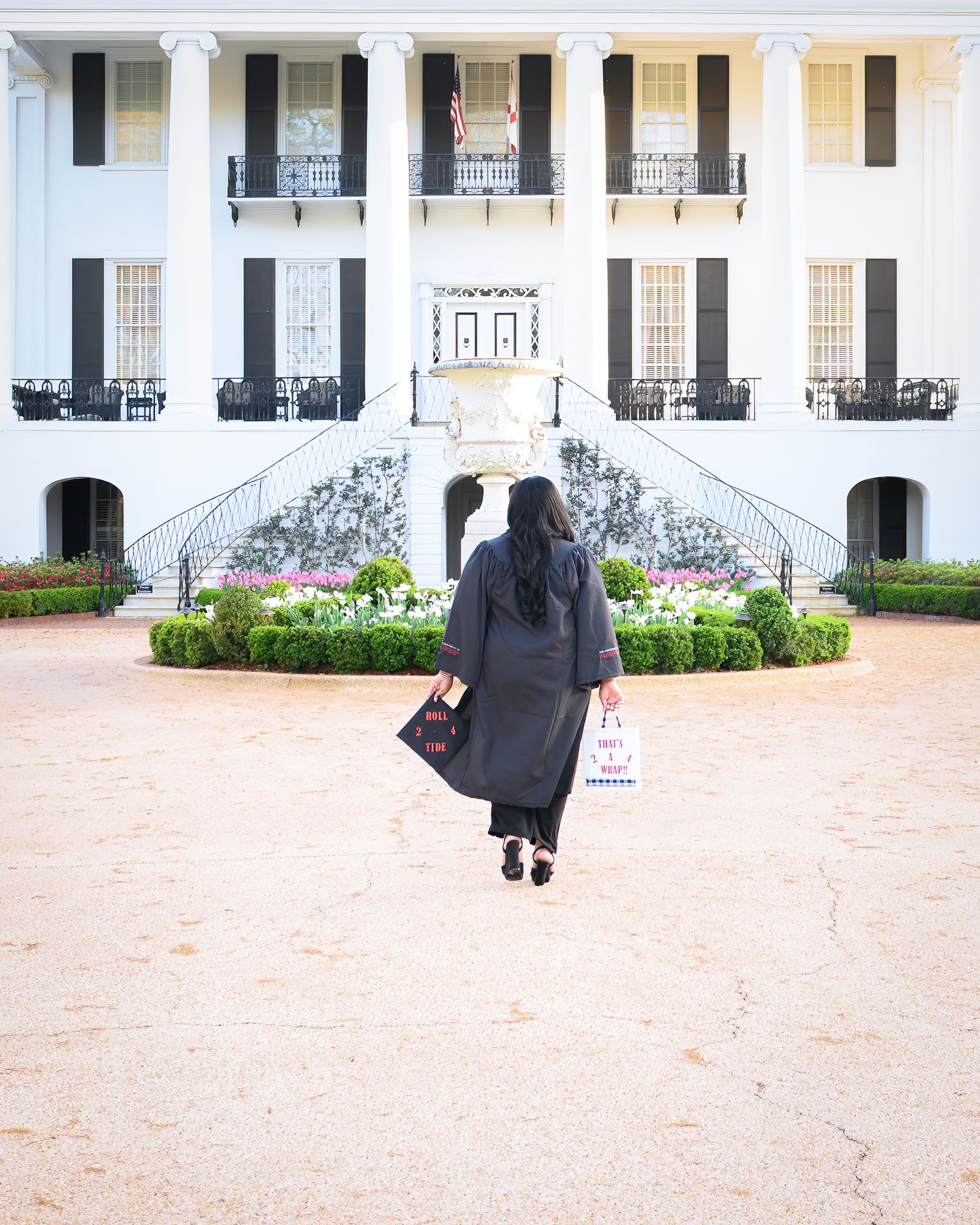 Graduate in black gown walks toward white columned building entrance.