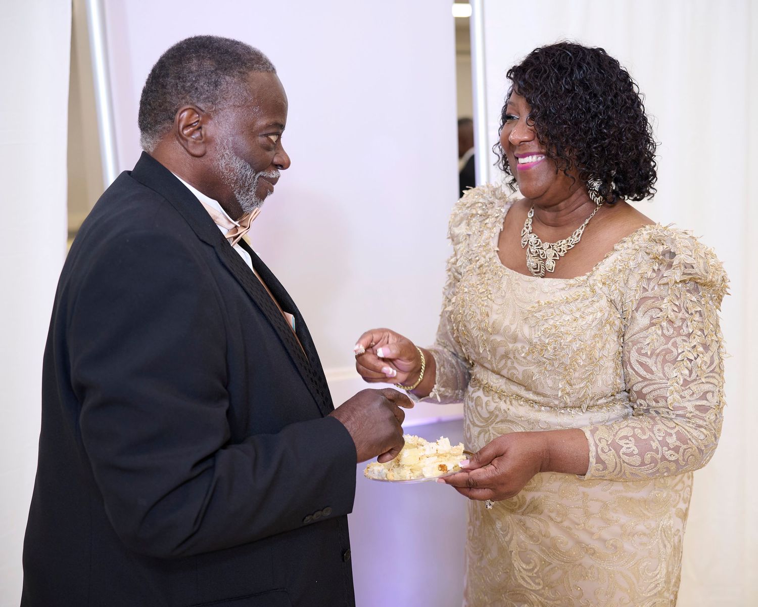 Couple sharing cake and sweet moments during their wedding reception against white backdrop.