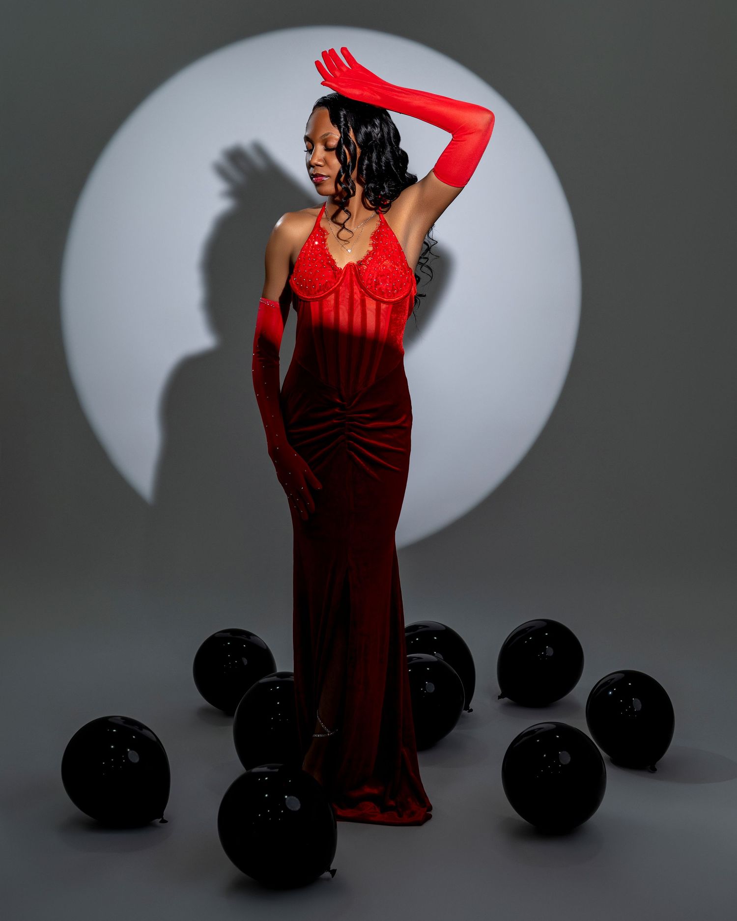 Model in red evening gown poses dramatically against circular spotlight with black balloons.