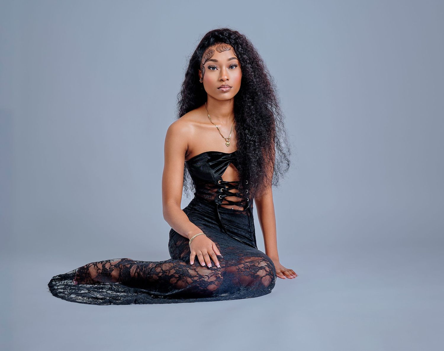 Model in black evening dress poses gracefully in studio against gray backdrop for fashion photoshoot series.