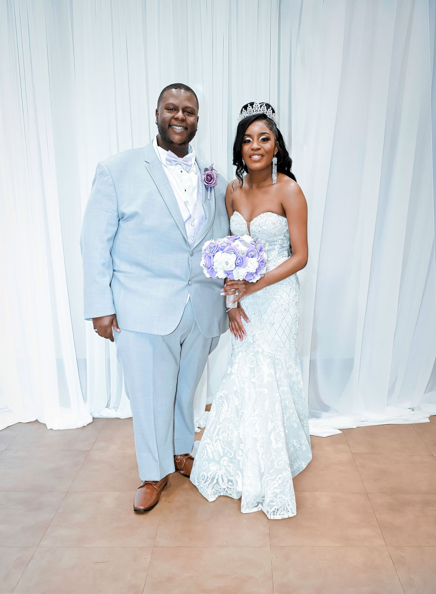 A couple in light blue and white formal attire poses with a purple bouquet.