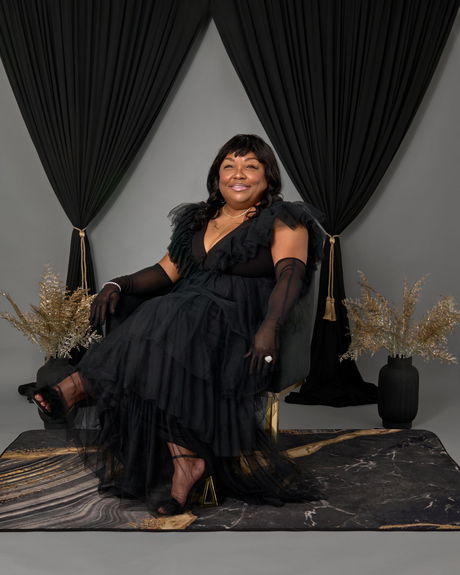 Glamorous portrait of a person in a black ruffled dress and gloves seated against dark curtains.