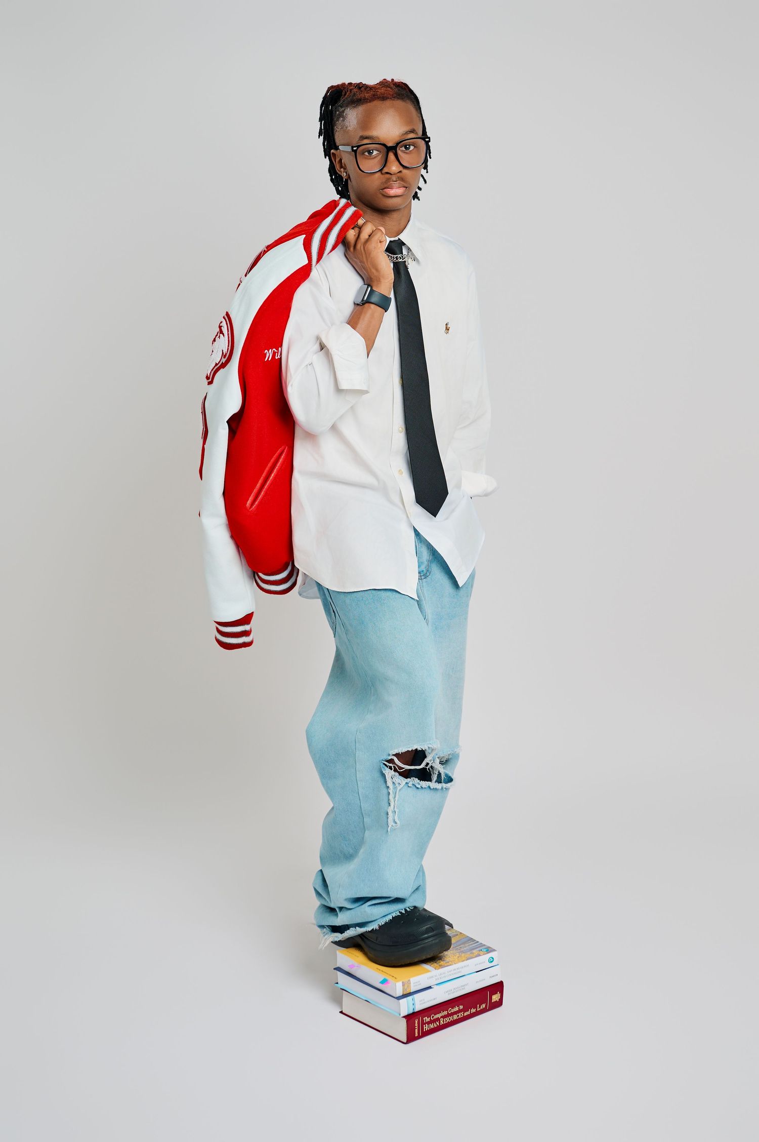 Person wearing light blue overalls with a red and white varsity jacket sits confidently on stacked books in a studio setting.