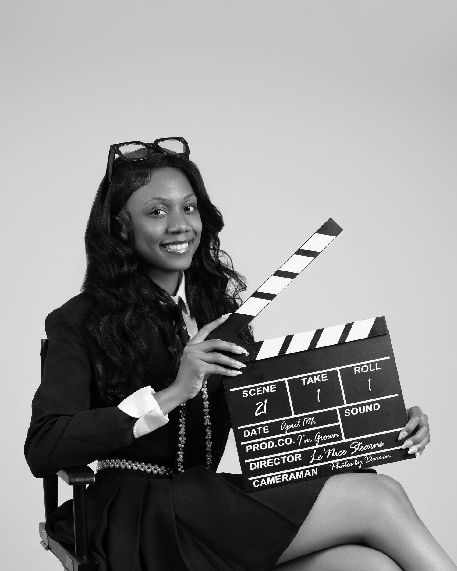 Black and white photo of a smiling woman holding a film clapperboard, wearing a blazer with sunglasses on her head.