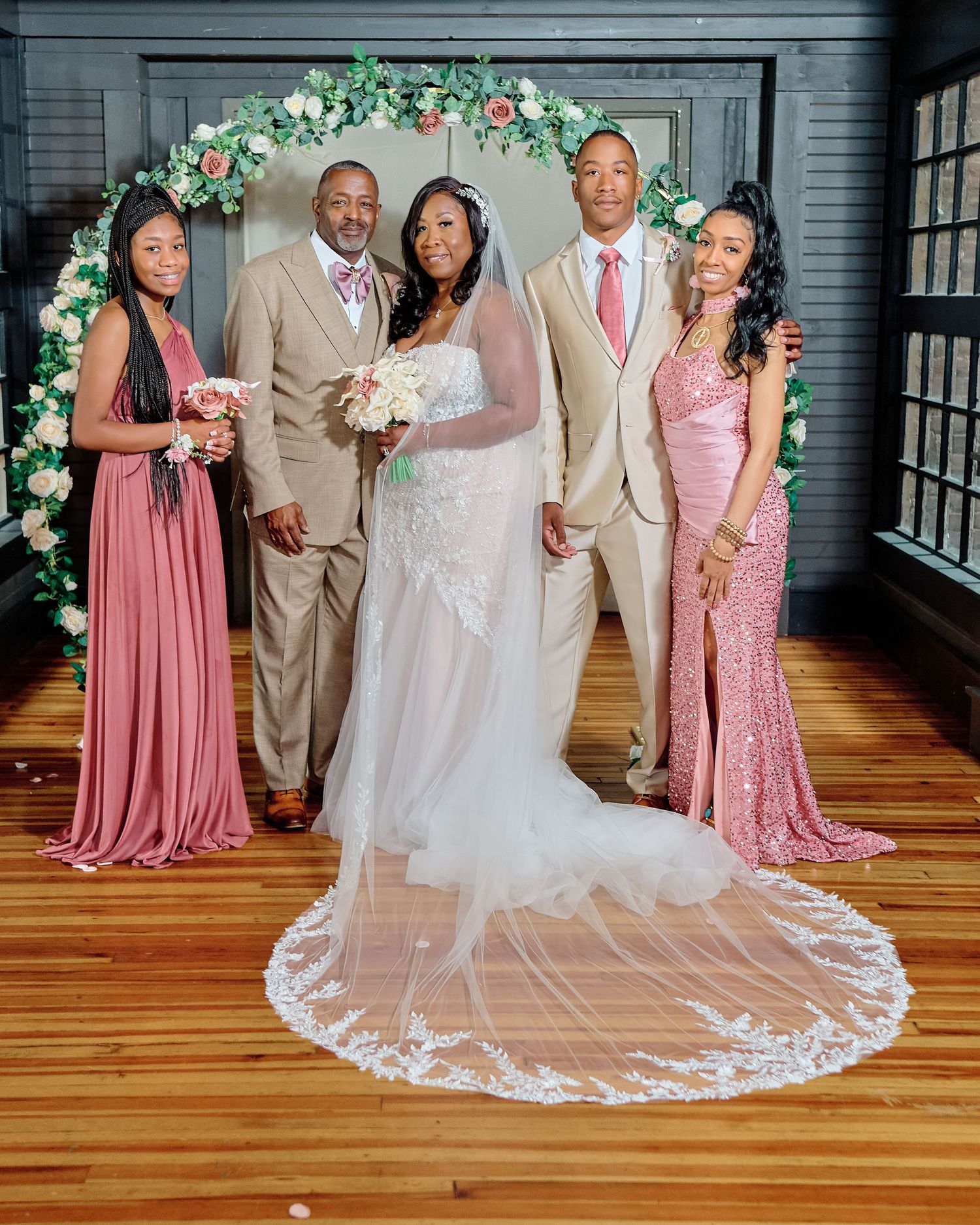Elegant wedding party poses in front of floral arch with bridesmaids in pink dresses and groomsmen in beige suits.