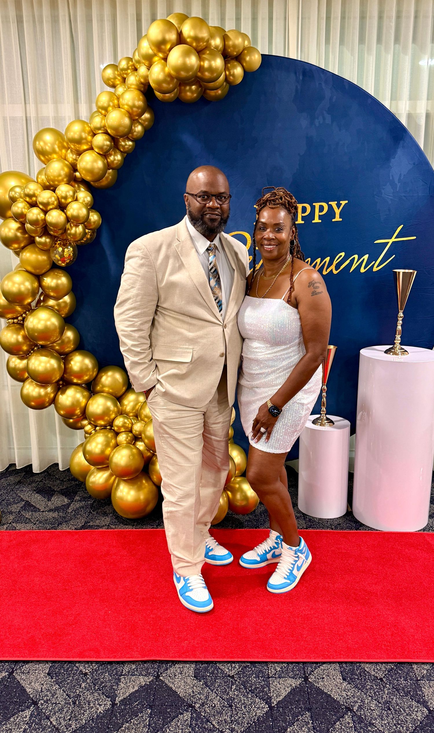 Couple poses on red carpet with gold balloon arch backdrop at formal event.