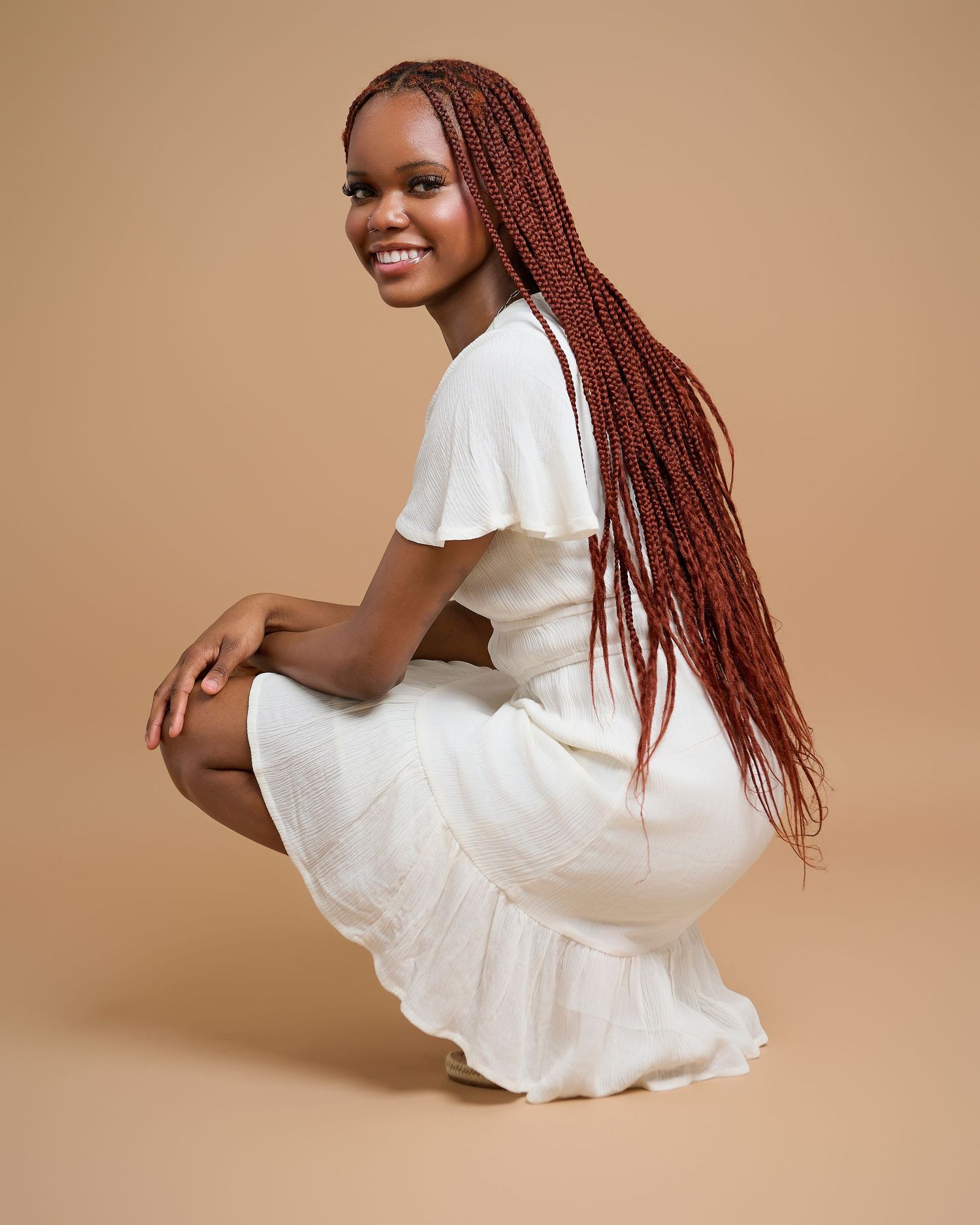 Model in white dress crouches while showcasing long red braids against beige background.