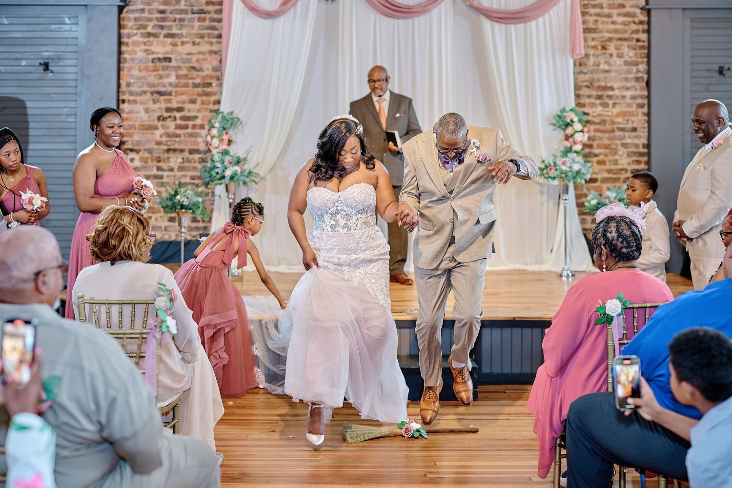 Wedding guests dance enthusiastically in a rustic reception venue with exposed brick walls and white draping.