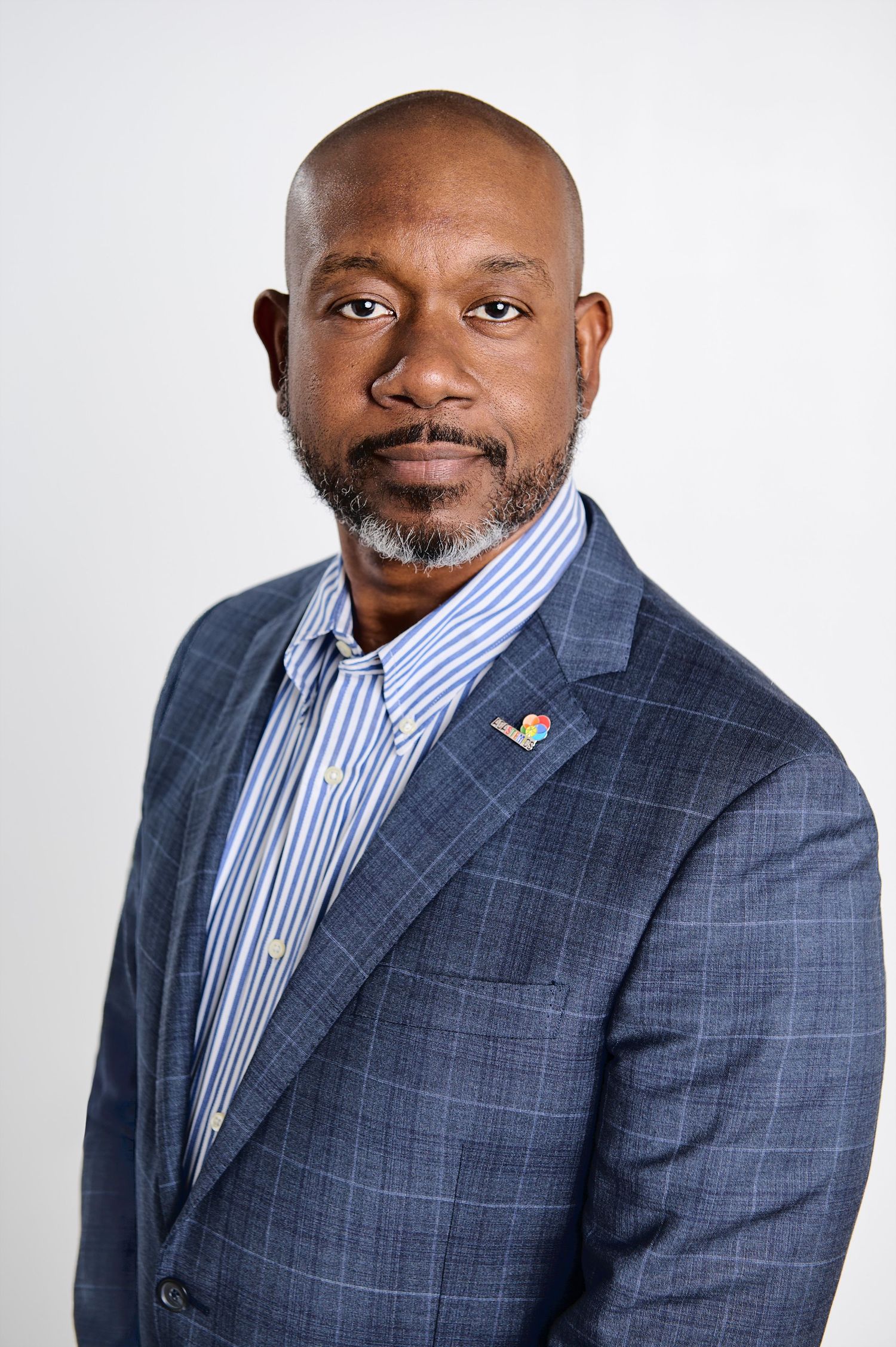 Professional headshot of person wearing blue plaid blazer and striped dress shirt against light background.
