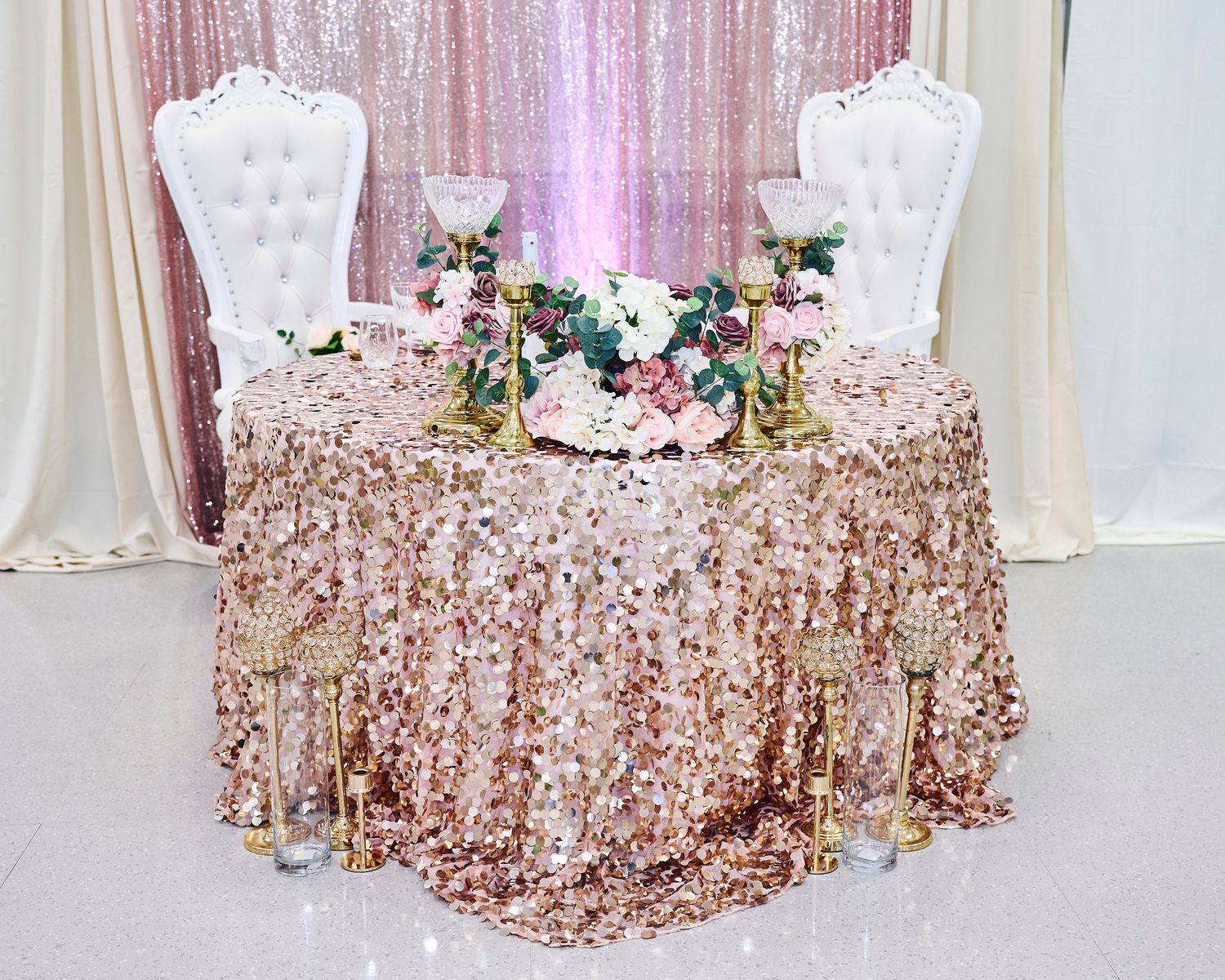 Elegant sweetheart table with rose gold sequin tablecloth, white throne chairs, and pink floral centerpieces at a wedding reception.