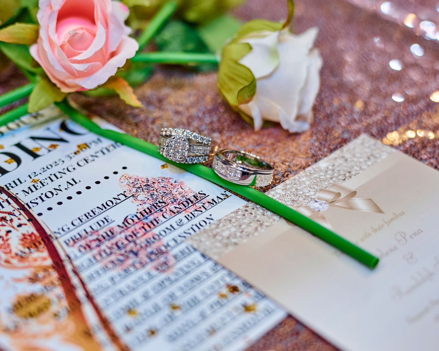 A wedding invitation lies on a glittery surface with pink roses and a diamond engagement ring displayed.