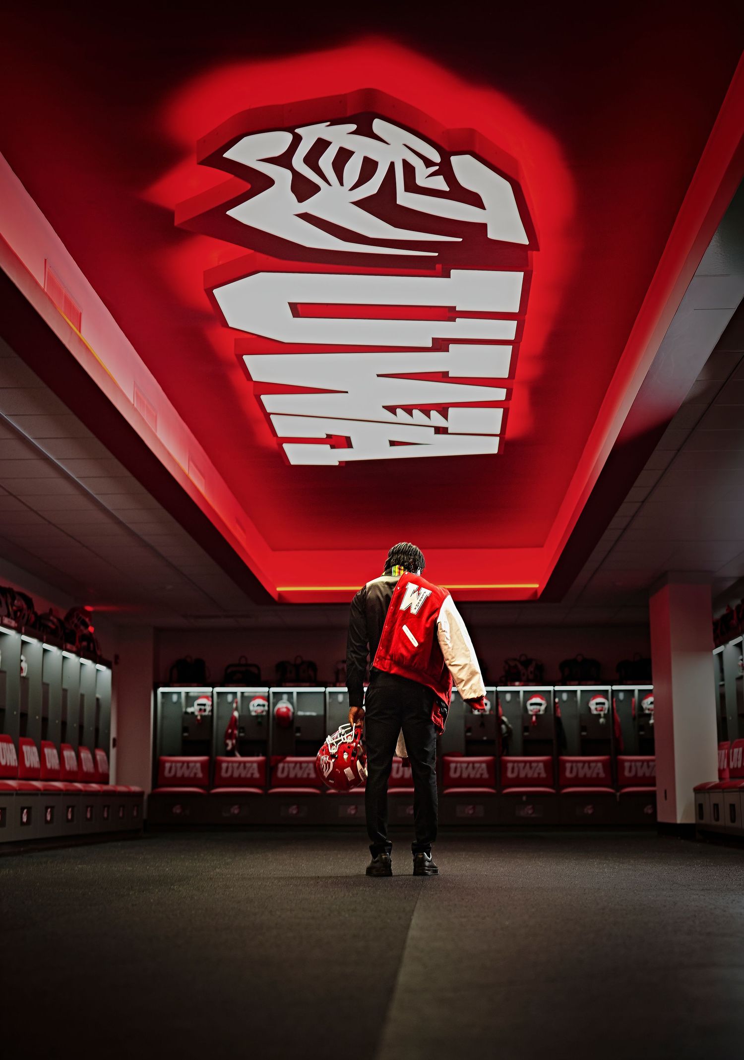 Person standing in red-lit sports locker room beneath glowing team logo on ceiling, wearing varsity jacket.